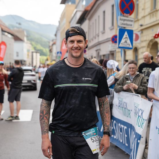 Ein Mann mit Tattoos und einem schwarzen Shirt, das Startnummer 5338 trägt, steht auf einer Straße während eines Marathons. Hinter ihm sind Zuschauer, ein Gebäude und ein Berg in der Ferne.