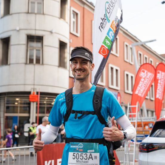 Ein Mann mit einem blauen Shirt und einem Visier-Cap nimmt an einem Marathon teil. Er hat die Nummer 5053A auf seinem Shirt. Hinter ihm befinden sich einige Gebäude, und Leute sind unterwegs.