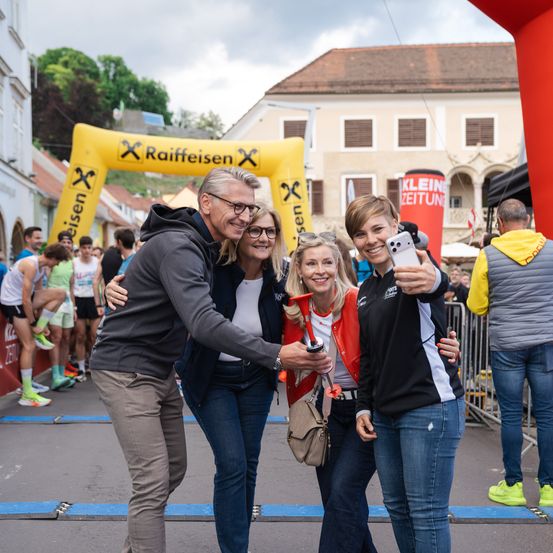 Vier Menschen posieren für ein Foto bei einem Laufevent vor einem gelben Bogen mit Raiffeisen-Branding. Dahinter sind weitere Läufer und Zuschauer zu sehen. Der Himmel ist bewölkt.