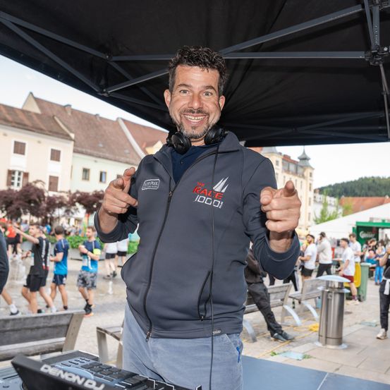 Ein Mann steht unter einem Zelt, lächelt und gestikuliert mit den Händen. Er trägt eine schwarze Kapuzenjacke mit 'The Race 1000' Schriftzug. Hinter ihm laufen Leute auf einer Straße mit Gebäuden und Bäumen.
