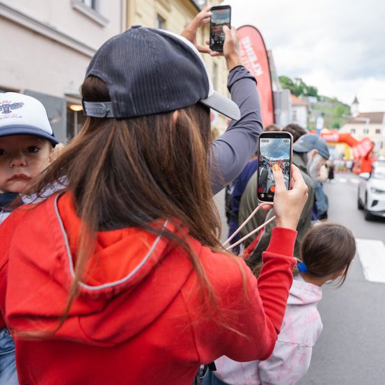 Eine Frau in einer roten Kapuzenjacke hält ein Handy und macht ein Foto von einem Straßenrennen. Ein Mann und ein Kind hinter ihr machen ebenfalls Fotos, mit Gebäuden und einem roten Banner im Hintergrund.