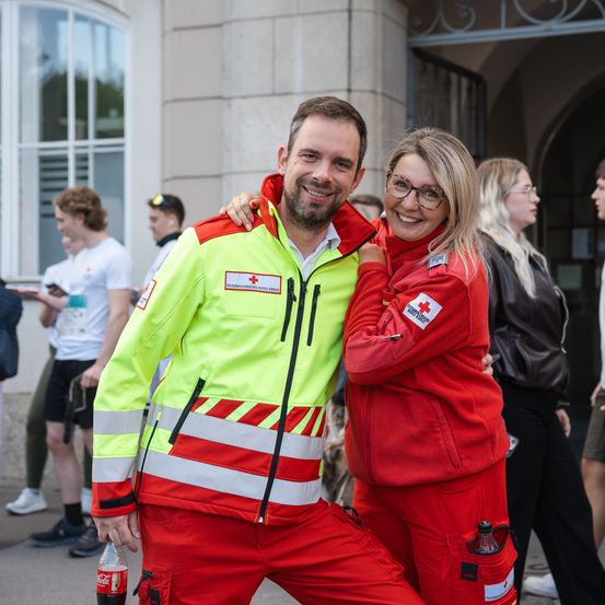 Ein Mann und eine Frau in roten und gelben Erste-Hilfe-Jacken lächeln für die Kamera, stehen vor einem Gebäude mit anderen Menschen im Hintergrund.