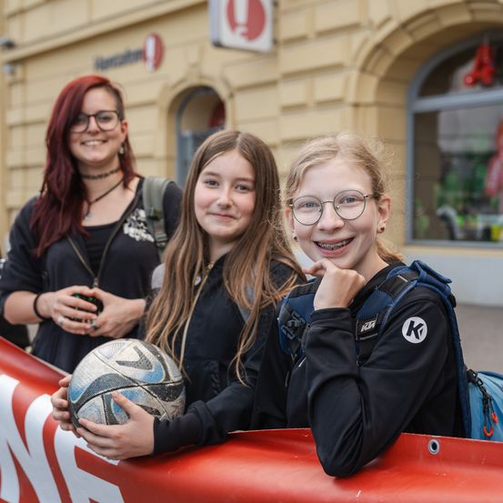 Drei junge Frauen stehen vor einem Gebäude mit einem Banner. Zwei halten einen Fußball.