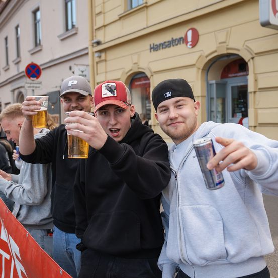 Drei Männer jubeln mit Getränken auf einer überfüllten Straße. Einer hält ein Bier und die anderen beiden haben Dosen Limonade. Sie stehen vor einem Gebäude mit einem Schild, das 'Hansaton' besagt.
