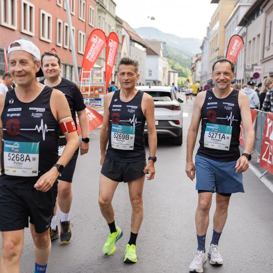 Vier Männer in schwarzen Tank-Tops und Shorts laufen einen Marathon. Sie haben Nummern auf ihren Trikots, und einer trägt ein rotes Armband. Ein weißes Auto und Gebäude sind im Hintergrund.