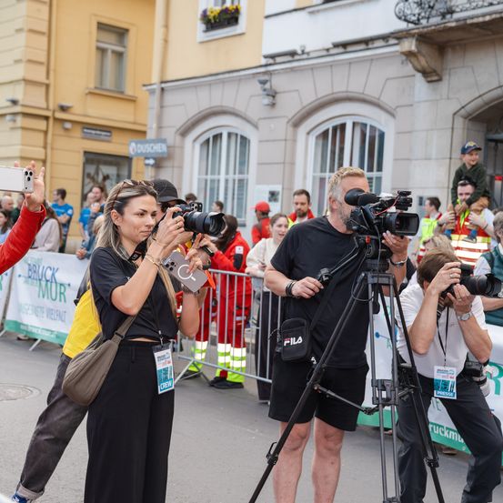 Mehrere Fotografen machen Fotos bei einer Outdoor-Veranstaltung, mit einer Frau und einem Mann mit einer Kamera und einem Stativ im Vordergrund. Dahinter versammelt sich eine Menschenmenge hinter einem Metallzaun.
