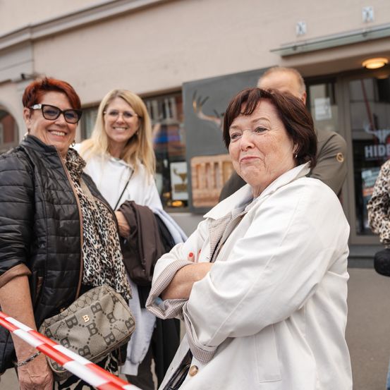 Eine Gruppe von Frauen steht vor einem Gebäude mit einem Hirschbild. Eine Frau verschränkt die Arme, während andere lächeln. Vor ihnen befindet sich ein Absperrband.