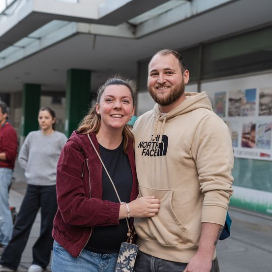 Ein Mann und eine Frau stehen lächelnd für ein Foto auf einer Stadtstraße. Der Mann trägt eine beigefarbene North Face-Kapuzenjacke und die Frau einen rotbraunen Blazer. Im Hintergrund gehen zwei Personen vorbei.