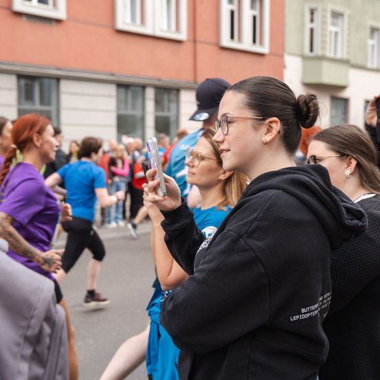 Eine Frau macht ein Foto von Läufern bei einem Straßenrennen. Zuschauer jubeln im Hintergrund, und Gebäude säumen die Straße.