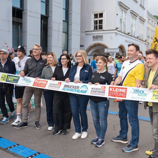 Eine Gruppe von Menschen steht hinter einem Banner für einen Marathon und lächelt für die Kamera. Sie halten ein langes Band mit Sponsorennamen. Gebäude sind im Hintergrund sichtbar.