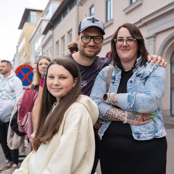 Eine Familie aus drei Personen steht vor einem Gebäude mit einem Gebäudeschild im Hintergrund. Der Mann trägt einen Hut und eine Brille, und die Frau hat ein Tattoo am Arm. Das Mädchen hat lange Haare und lächelt.