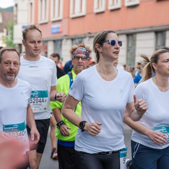 Eine Gruppe von Läufern nimmt an einem Marathon teil. Eine Frau vorne trägt eine Sonnenbrille und ein weißes T-Shirt. Ein anderer Läufer hinter ihr trägt eine gelbe Weste. Gebäude sind im Hintergrund sichtbar.