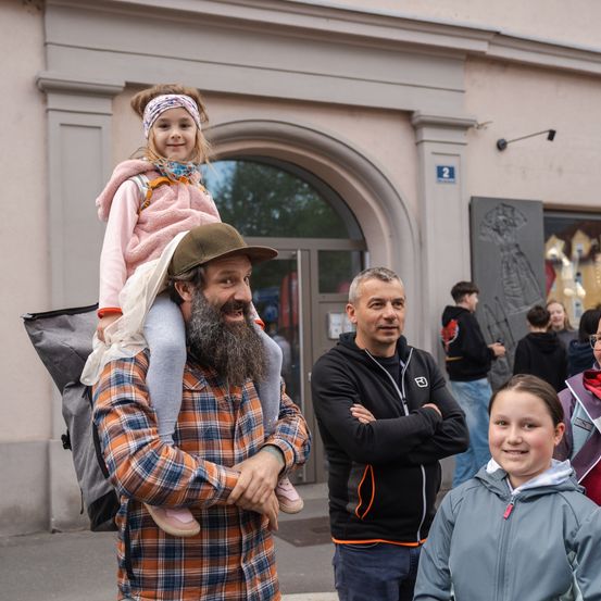 Ein Mann mit Bart trägt ein junges Mädchen auf seinen Schultern vor einem Gebäude, mit anderen Leuten, die herumstehen.