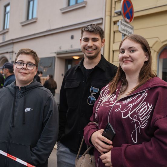 Drei Personen, zwei Männer und eine Frau, stehen lächelnd nebeneinander draußen, mit einem Gebäude, einem Schild und einer Barriere im Hintergrund.