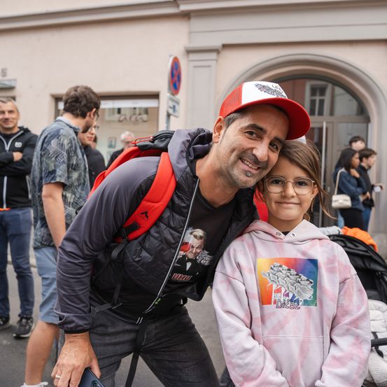 Ein Mann mit Rucksack und Kappe posiert mit einem lächelnden Mädchen in einer Tie-Dye-Hoodie auf einer belebten Straße. Im Hintergrund laufen Leute, und ein Kinderwagen steht in der Nähe.