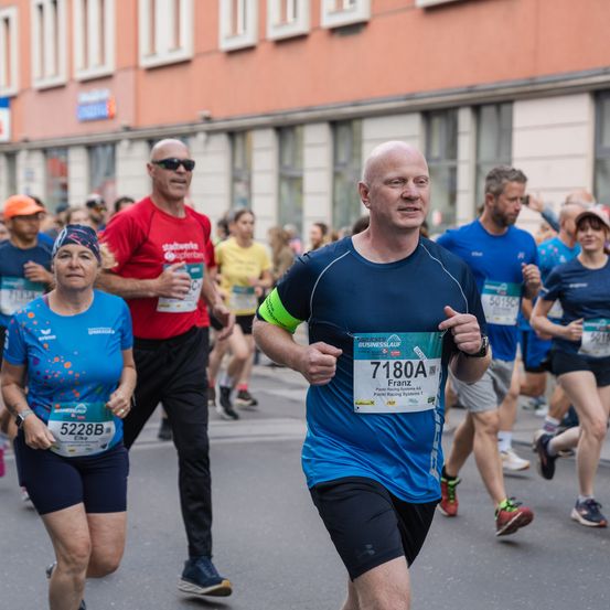 Eine Gruppe von Menschen nimmt an einem Marathon teil. Ein Mann mit der Nummer 71804 führt, gefolgt von einer Frau und einem weiteren Mann. Sie laufen mitten auf der Straße. Dahinter sind weitere Teilnehmer zu sehen. Gebäude säumen die Straße auf beiden Seiten.