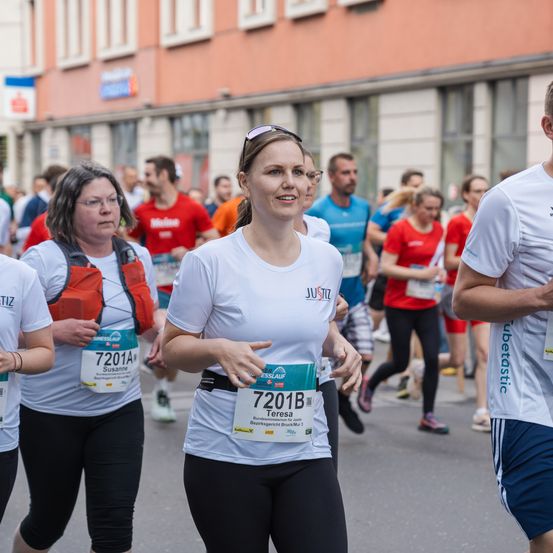 Eine Gruppe von Marathonläufern mit weißen Shirts läuft auf der Straße. Eine Frau vorne lächelt. Dahinter laufen Menschen, und ein Gebäude ist an der Seite.