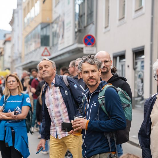 Eine Gruppe von Menschen geht auf einer Straße mit Gebäuden im Hintergrund. Ein Mann hält ein Getränk, während ein anderer ein Telefon hält. Es gibt Schilder und eine Menschenmenge.