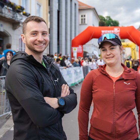 Ein lächelnder Mann und eine Frau stehen gemeinsam draußen in der Nähe einer Marathon-Ziellinie mit Zuschauern im Hintergrund.