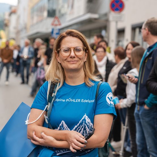Eine Frau mit Brille lächelt, während sie auf der Straße mit einer Menschenmenge steht. Sie trägt ein blaues T-Shirt mit dem Alpine Bohler Edelstal Logo.