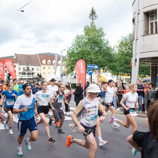 Eine Gruppe von Läufern nimmt an einem Marathon auf einer Stadtstraße teil, mit Gebäuden und Zuschauern im Hintergrund. Rote Banner und Straßenschilder sind sichtbar.