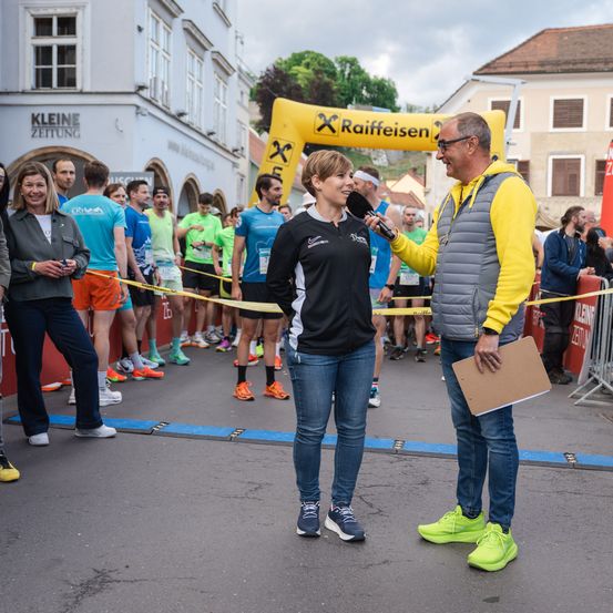 Eine Frau wird von einem Mann bei einem Marathon interviewt. Sie stehen vor Läufern und einem Gebäude mit dem Schriftzug 'Kleine Zeitung'.