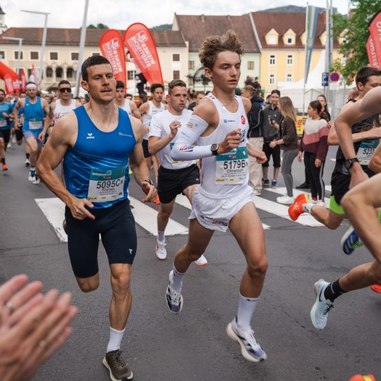 Mehrere Läufer nehmen an einem Marathon teil. Ein Läufer in Blau führt, gefolgt von einem anderen in Weiß. Zuschauer beobachten von den Seitenlinien. Gebäude säumen die Straße, mit Fahnen und Schildern im Hintergrund.