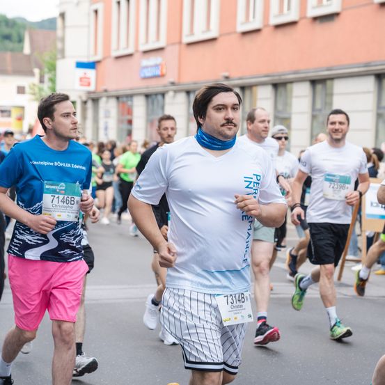 Eine Gruppe von Marathonläufern nimmt an einem Rennen teil. Ein Läufer trägt ein weißes T-Shirt mit einer blauen Bandana, während ein anderer Läufer ein blaues T-Shirt mit einem weißen und blauen Design trägt. Im Hintergrund sind Gebäude und Menschen zu sehen.