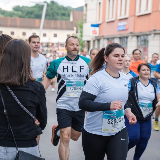 Eine Gruppe von Menschen läuft in einem Marathon. Eine Frau mit der Nummer 5200C schaut nach rechts. Hinter ihr läuft ein Mann mit einem grünen und weißen Trikot mit der Nummer 72058.