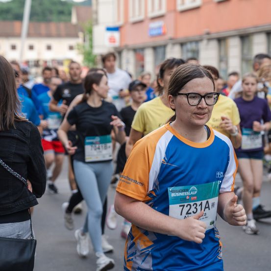 Ein Marathonlauf ist im Gange. Die Teilnehmer tragen nummerierte Startnummern. Ein Mädchen mit Brille läuft vorne und lächelt, während andere hinterherlaufen. Im Hintergrund sind Gebäude und Bäume zu sehen.