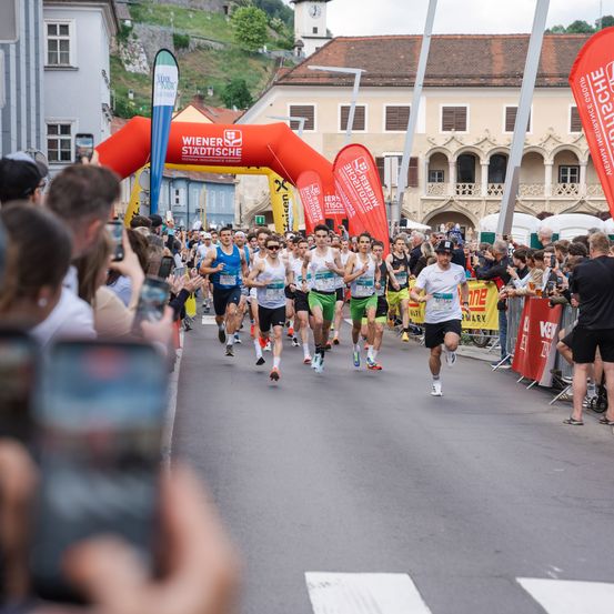Ein Marathonrennen mit Läufern in verschiedenen Farben und Nummern ist in einer Stadt im Gange. Zuschauer beobachten von den Seitenlinien.