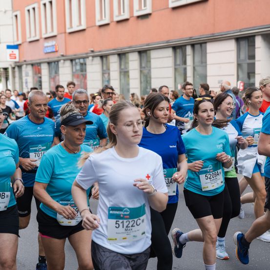 Läufer nehmen an einem Marathon teil, tragen Nummern, Brillen und Mützen. Ein Gebäude mit Fenstern ist hinter ihnen.