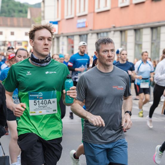 Ein Läufer in einem grünen Shirt mit der Nummer 5140A läuft einen Marathon mit einer Wasserflasche in der Hand, während ein älterer Mann in einem grauen Shirt zuschaut.