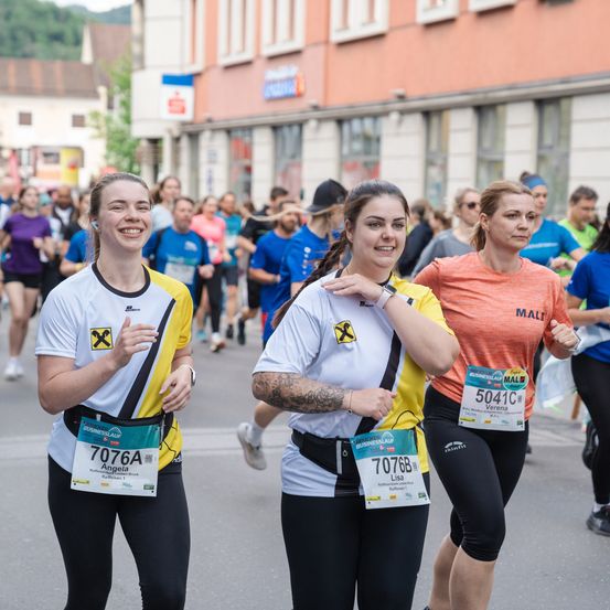 Läuferinnen bei einem Marathon mit Nummern auf ihren Trikots. Sie lächeln, und eine von ihnen hat die Startnummer 7076A. Das Gebäude im Hintergrund hat viele Fenster.