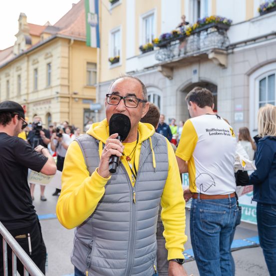 Ein Mann in einem gelben Sweatshirt spricht in ein Mikrofon bei einer Veranstaltung im Freien, mit Gebäuden und einer Menschenmenge im Hintergrund.