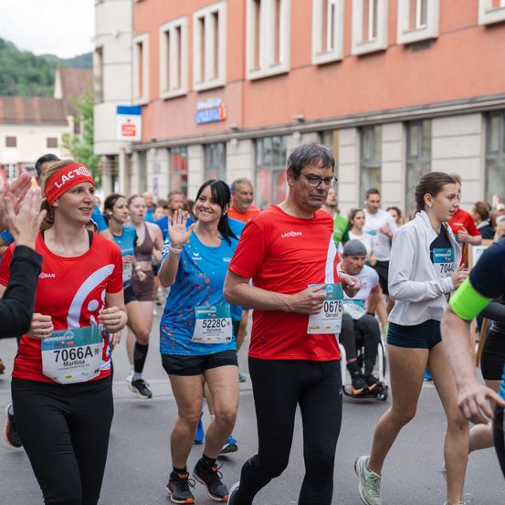 Teilnehmer eines Marathons laufen eine Straße hinunter, mit Gebäuden und Grünflächen im Hintergrund. Ein Mann in Rot führt das Rennen an.