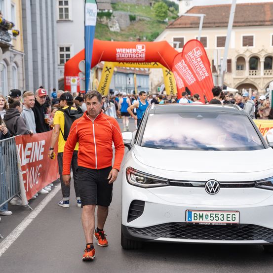 Ein älterer Mann in einer orangefarbenen Jacke geht an einem weißen Elektroauto von Volkswagen auf einer Stadtstraße vorbei, mit einer Menge von Läufern und Bannern im Hintergrund.