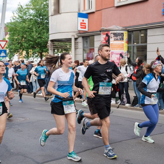 Läufer nehmen an einem Marathon teil, wobei mehrere Teilnehmer im Hintergrund zu sehen sind. Der Mann vorne hat die Nummer 5306C, die Frau neben ihm 7307B. Zuschauer jubeln von der Seitenlinie.