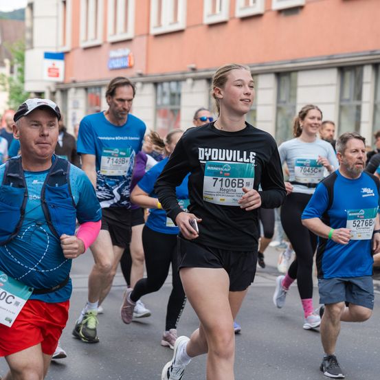 Läufer nehmen an einem Marathon teil und tragen nummerierte Startnummern. Eine Frau in der Mitte trägt ein schwarzes Shirt mit Dyduville und 7106B. Dahinter ein Mann in einem blauen Shirt mit der Nummer 7149B. Im Hintergrund sind Gebäude und ein Banner zu sehen.