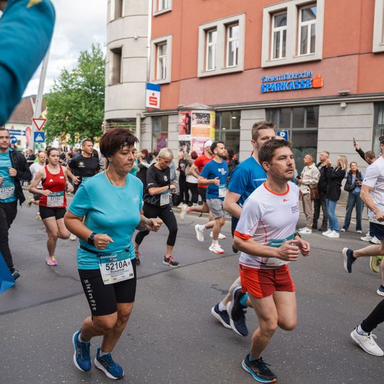 Ein Marathon findet in der Stadt statt, bei dem mehrere Läufer Nummern tragen. Zuschauer beobachten von der Seite. Ein Gebäude mit dem Schriftzug 'Steiermarkische Sparkasse' ist im Hintergrund.
