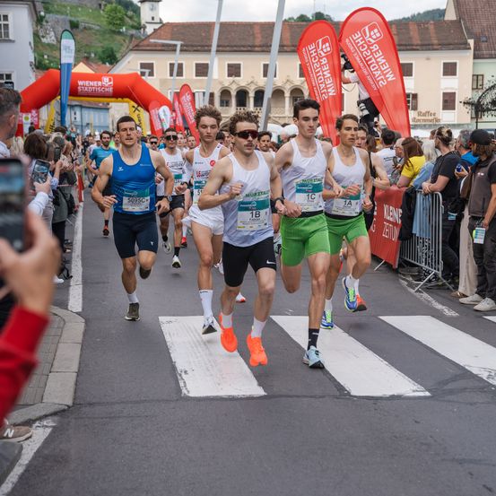 Ein Marathonlauf findet in einer Stadtstraße statt, bei dem mehrere Läufer mit nummerierten Trikots und Turnschuhen unterwegs sind. Zuschauer jubeln von den Rändern.