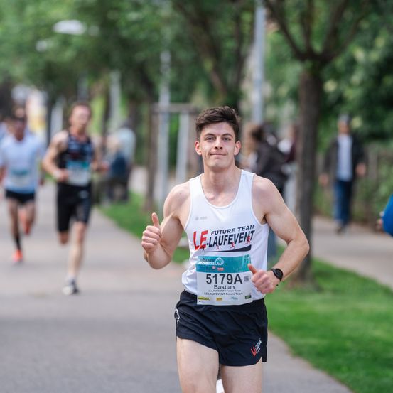 Ein Mann mit der Nummer 5179A auf seinem Trikot läuft bei einem Marathon. Er gibt mit der rechten Hand einen Daumen hoch. Hinter ihm nehmen auch andere Läufer am Marathon teil.