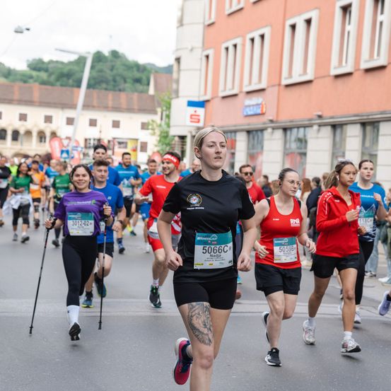Eine Gruppe von Läufern mit Startnummern nimmt an einem Marathon teil. Die Frau vorne hat die Nummer 5066. Im Hintergrund befinden sich Gebäude und Grünflächen.