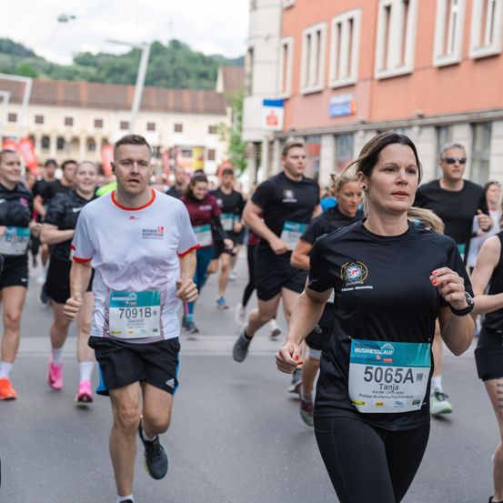 Teilnehmer eines Marathonlaufs, mit einem Mann und einer Frau in Führung, umgeben von anderen im Hintergrund. Im Hintergrund sind Gebäude zu sehen.