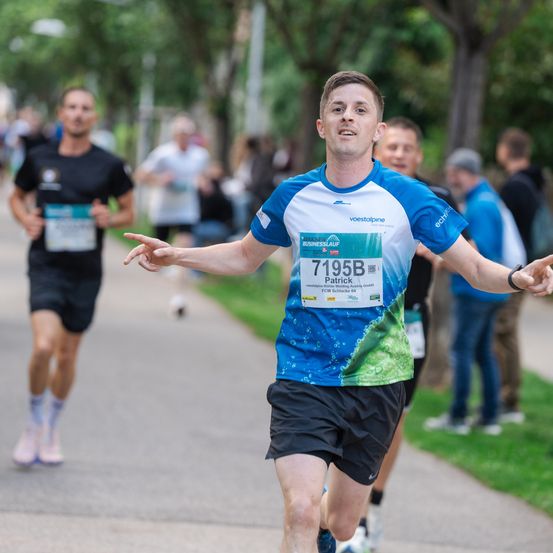 Ein Marathonläufer, Patrick, trägt ein blaues und weißes Trikot mit der Nummer 7195B und läuft im Vordergrund. Hinter ihm sind andere Läufer und Zuschauer zu sehen, mit Bäumen und Gras im Hintergrund.