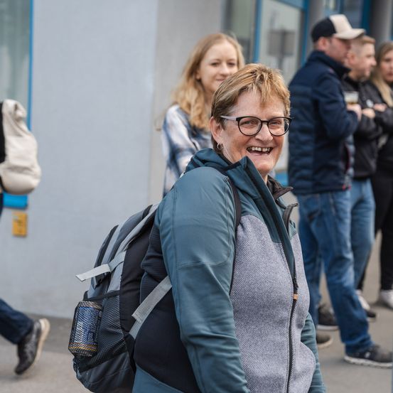 Eine Frau mit einem Rucksack lächelt vor einem Gebäude, während andere im Hintergrund stehen.