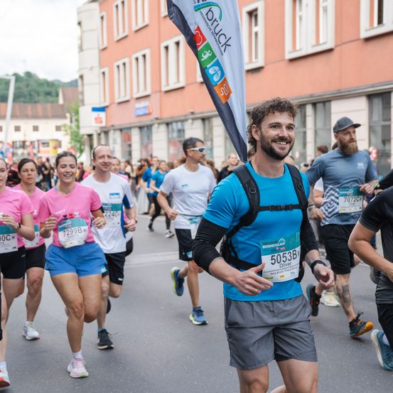 Läufer nehmen an einem Marathon teil. Sie tragen nummerierte Startnummern und tragen Flaggen. Der Läufer vorne trägt einen Rucksack. Gebäude säumen die Straße.