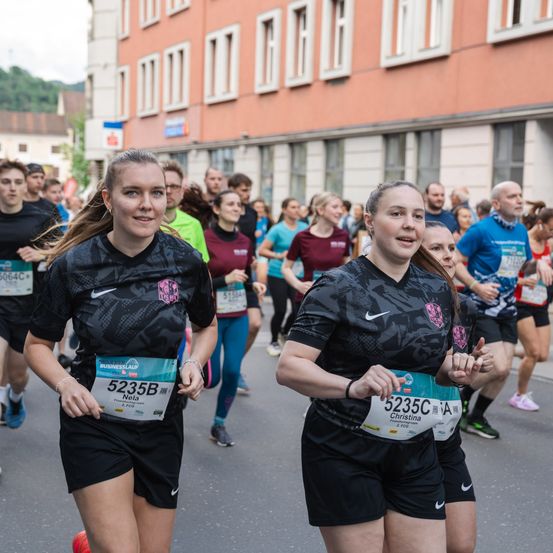 Läufer nehmen an einem Marathon teil und tragen Nummern. Eine Frau vorne lächelt, und eine andere Frau hinter ihr sieht voraus. Sie sind von anderen Läufern umgeben, mit einem Gebäude im Hintergrund.