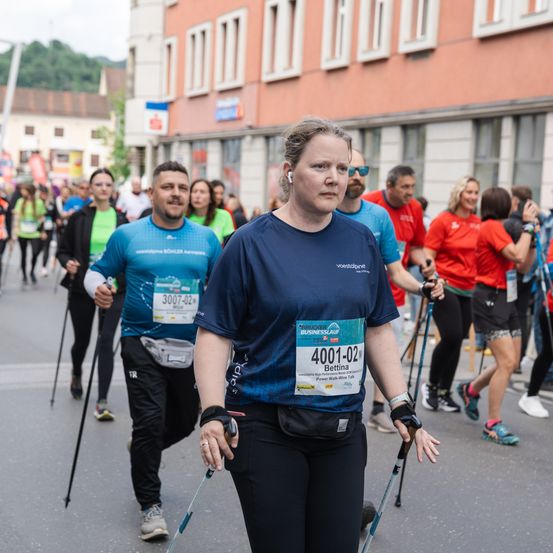 Eine Frau und zwei Männer nehmen an einem Marathon teil, tragen Startnummern 4001-02, 3007-02 und 3006-02. Sie laufen mit Stöcken, tragen blaue Shirts und sind von einer Menge umgeben. Gebäude und Bäume sind im Hintergrund.