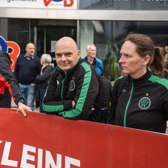 Ein Mann und eine Frau stehen vor einem roten Banner mit dem Wort Kleine darauf. Sie tragen Jacken mit demselben Logo. Dahinter stehen mehrere Leute vor einem Gebäude.